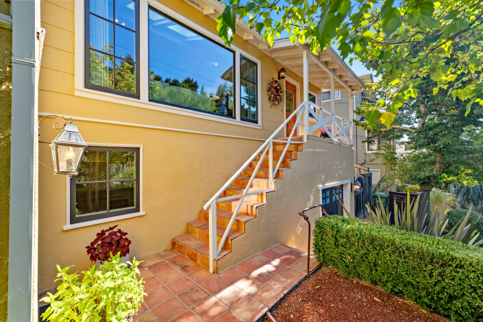 A yellow house at 260 Clorinda Avenue San Rafael features large windows, terracotta-tiled steps leading to a white-railed porch, green shrubs, potted plants, and a tree providing shade in the foreground.