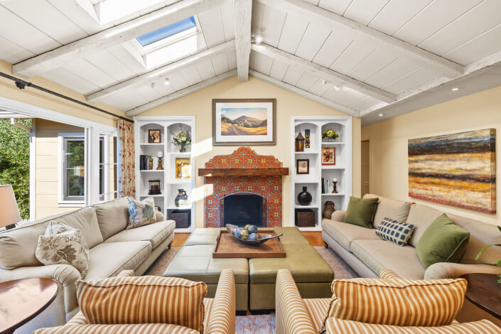 A bright living room with two beige sofas, striped armchairs, a patterned ottoman, a colorful tiled fireplace, built-in shelves, a large landscape painting above the mantel, and a skylight in the white beamed ceiling.