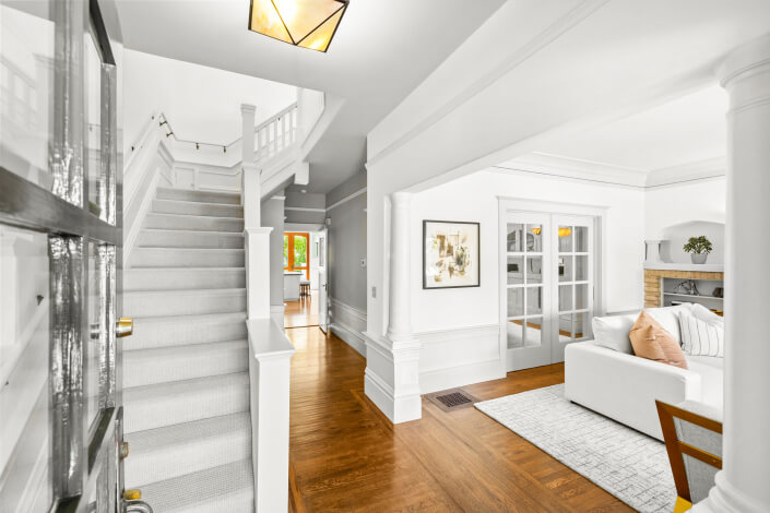 Bright, modern home interior with white walls and trim, light carpeted staircase on the left, hardwood floors, a cozy living room with a white sofa, and glass French doors leading to another room.