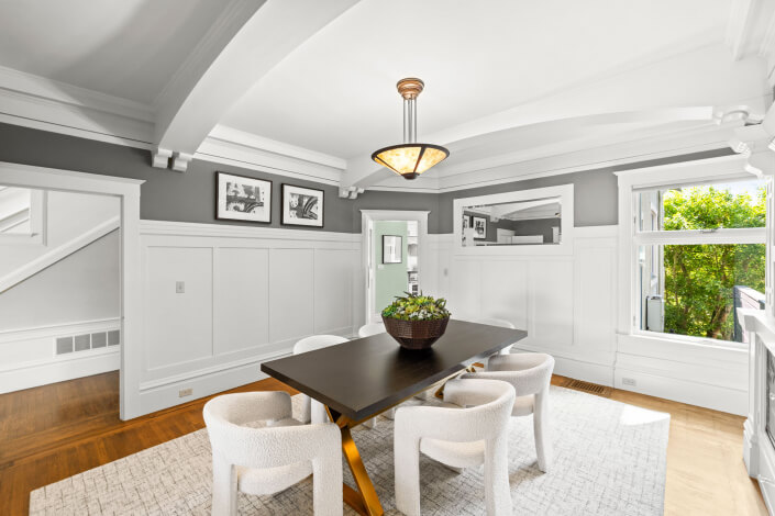 A modern dining room with a long dark table, white cushioned chairs, a basket of green plants as centerpiece, white built-in cabinets, gray walls, and black-and-white photos on the walls. A chandelier hangs above.