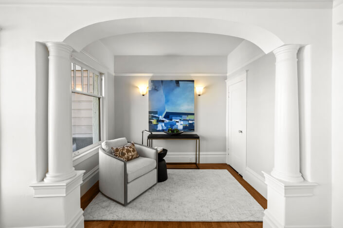 A small, elegant nook with white walls and columns, featuring a gray armchair, a side table, a console table, and a large blue abstract painting under two wall sconces. A light rug covers the wooden floor.