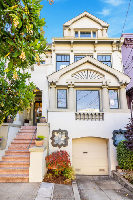 A cream-colored, multi-story house at 739 10th Avenue San Francisco features large windows, decorative architectural details, and a garage. Steps with potted plants lead to the entrance, with trees and shrubs framing the front.
