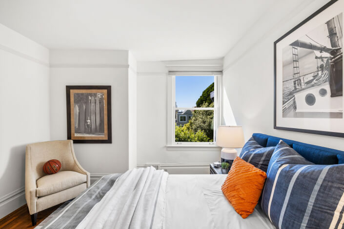 Bright bedroom with white walls, a neatly made bed with blue and orange pillows, a beige armchair with a decorative pillow, framed artwork, and a window showing trees and a building outside.