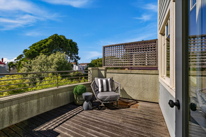 Sunny balcony with wooden flooring, a cushioned chair with striped pillows, a small side table, and a potted plant. Trees and buildings are visible in the background under a clear blue sky.