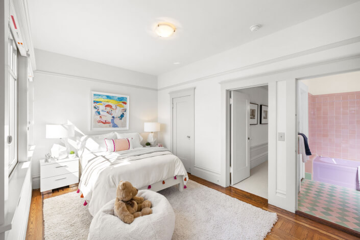 Bright bedroom with a bed, white bedding, two nightstands, and lamps. A colorful painting hangs above the bed. A stuffed bear sits on a beanbag chair, and a pink-tiled bathroom is visible through an open door.