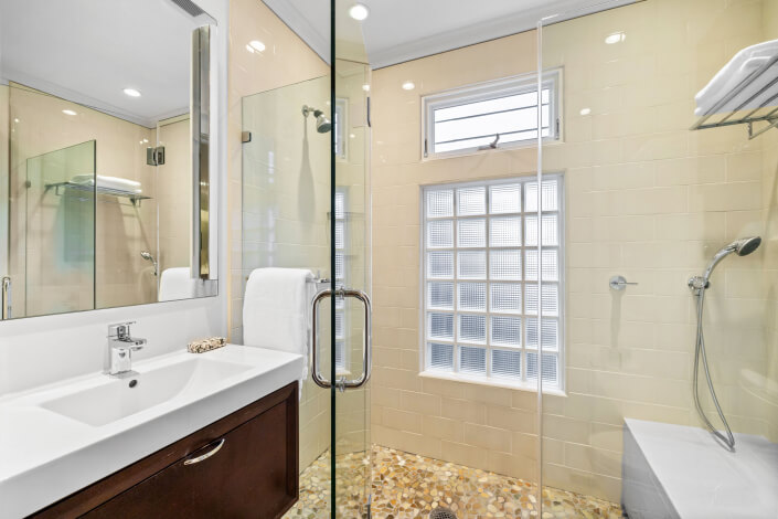 Modern bathroom with glass shower enclosure, pebble tile floor, beige tiled walls, wall-mounted showerhead, rectangular window with glass blocks, white sink, and dark wood vanity with a towel hanging on the wall.