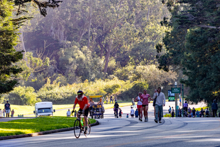 People enjoy a sunny day in a park; some are walking, others cycling or riding a surrey bike, with green trees and grass in the background and sunlight filtering through the foliage.