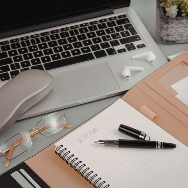 An open laptop, eyeglasses, a notebook with “to do list” written on it, a pen, wireless earbuds, and a potted succulent sit on a desk, creating a tidy and organized workspace.