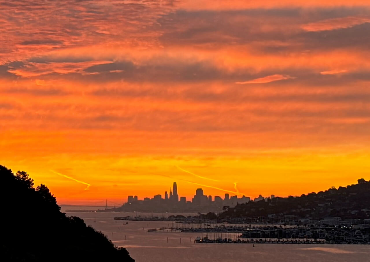 From De Silva Island: Sunrise over the San Francisco skyline, with orange and yellow hues in the sky, silhouetted buildings, the bay, and hills framing the city.