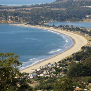 Stinson Beach, California: Aerial view of a long, curving sandy beach bordered by blue water on one side and a coastal town with houses and green trees on the other, surrounded by hills and distant landscape.