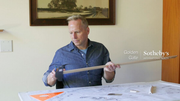 Thomas Henthorne sits at a drafting table with landscape plans The Golden Gate Sotheby’s International Realty logo appears in the top right corner. A framed landscape painting hangs on the wall behind him.