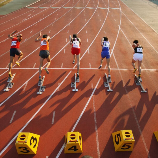 Five athletes are starting a race on a red track, each in a separate lane, pushing off from starting blocks, with numbered lane markers visible in the foreground.