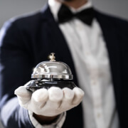 A person in a suit and bow tie, wearing a white glove, holds out a silver service bell on a black base, suggesting luxury service. The background is blurred.