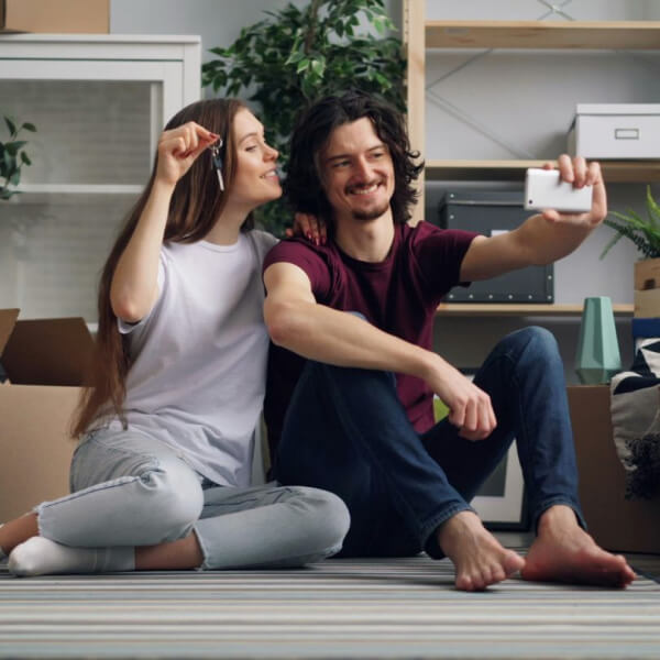 A smiling couple sits on the floor of a new home, holding keys and taking a selfie together. Cardboard boxes and a plant are visible in the background, suggesting they have just moved in.