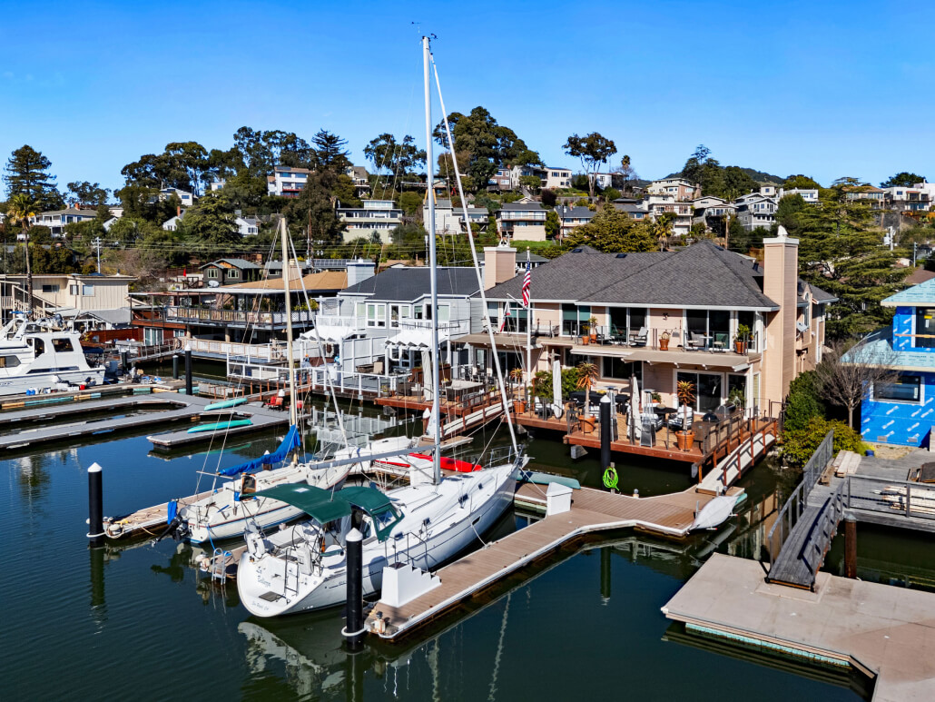 Two sailboats docked at a marina in front of waterfront houses with decks overlooking the water at 26 Mooring Road in San Rafael; hillside homes and trees rise in the background under a clear blue sky.