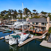 Two sailboats docked at a marina in front of waterfront houses with decks overlooking the water at 26 Mooring Road in San Rafael; hillside homes and trees rise in the background under a clear blue sky.