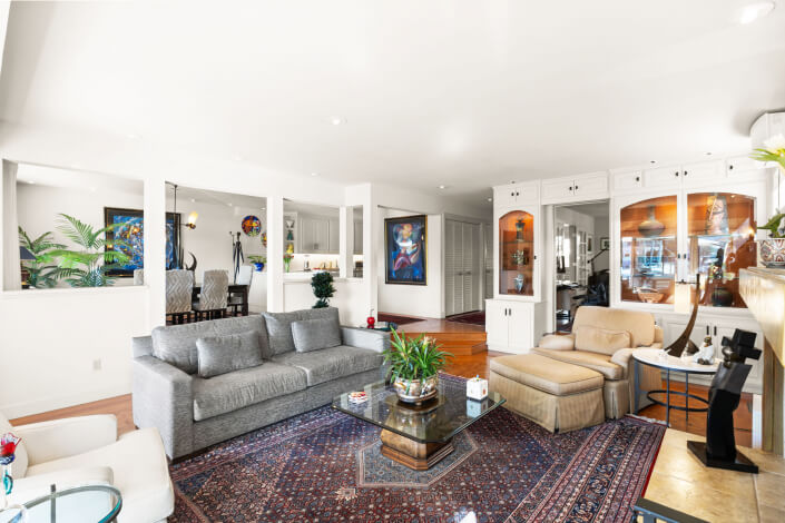 A bright, spacious living room features a gray sofa, beige armchairs, a glass coffee table with plants, built-in white shelves, and a patterned rug. Artwork and plants decorate the space, with a dining area in the background.