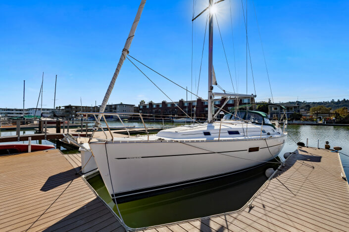A white sailboat docked at a marina on a sunny day, with calm water and waterfront buildings in the background. The sun shines brightly behind the mast.