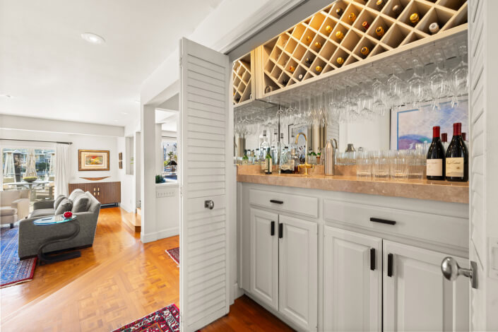 A home bar with white cabinets, a marble countertop, wine bottles, glasses, and liquor bottles. Open louvered doors reveal a wine rack above. A living room with modern furniture is visible in the background.