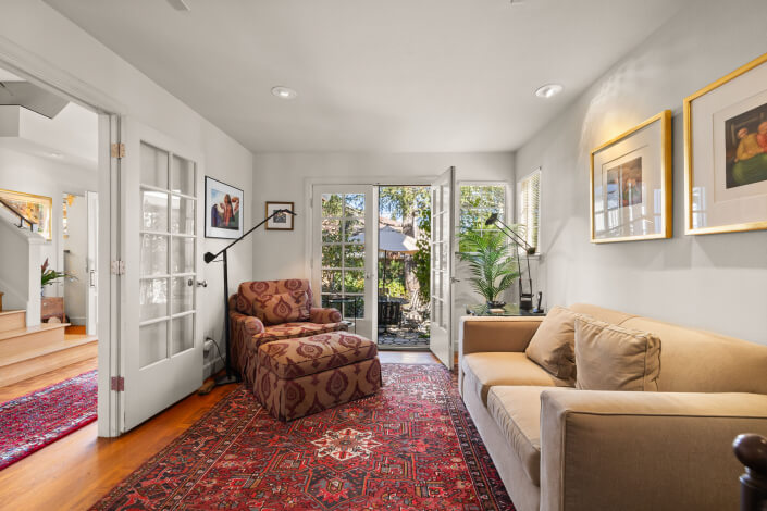 A cozy living room with a beige sofa, patterned armchair, red rug, and framed art on white walls. Glass doors open to a garden patio, letting in natural light. A plant and floor lamps add warmth to the inviting space.