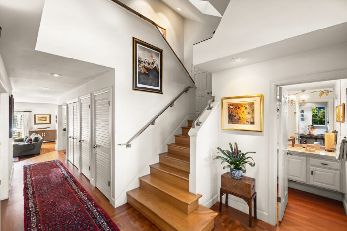 A bright hallway with wooden floors and stairs, white walls, colorful artwork, a long red rug, and a small table with a plant. Doors line the left wall and powder room is visible to the right.