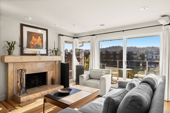 A bright living room at 26 Mooring Road in San Rafael features a stone fireplace with artwork above, gray sofas, a wooden coffee table, potted plants, and large windows overlooking the canal and neighboring houses.