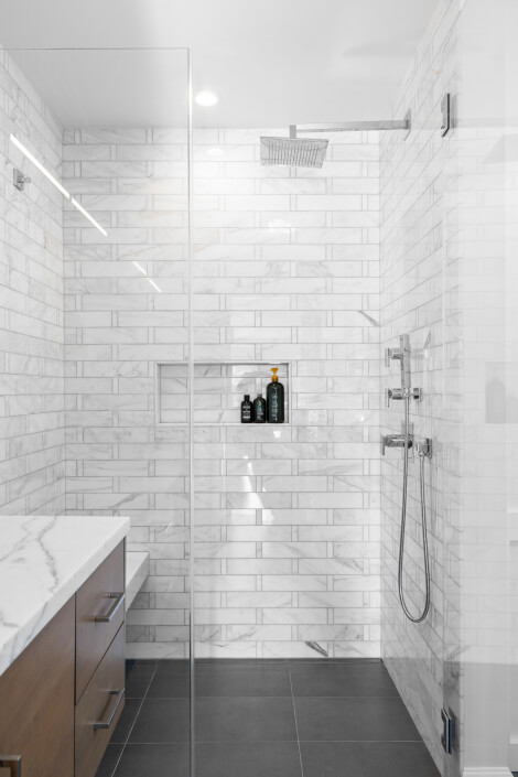 Modern bathroom with white marble subway tile shower, built-in wall niche holding toiletries, overhead rainfall showerhead, handheld shower, glass partition, dark gray floor tiles, and a wood vanity with a marble countertop.