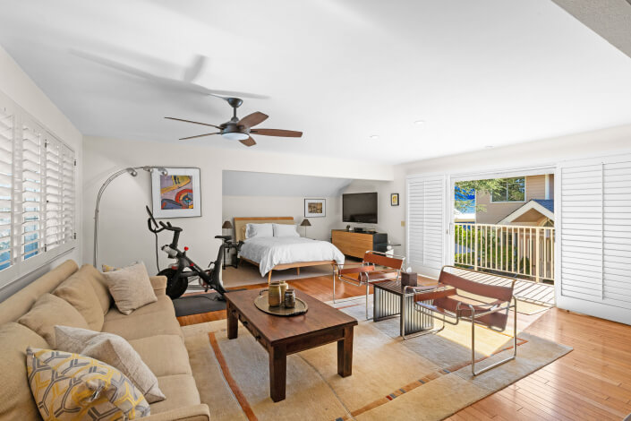 A bright, modern bedroom with a bed, TV, exercise bike, beige sofa, coffee table, and chairs. Large windows with white shutters let in natural light, and a balcony is visible outside.