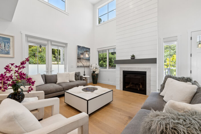 Bright, modern living room at 31 Gold Hill Grade San Rafael with high ceilings, large windows, two gray sofas, white armchairs, a square coffee table, fireplace with white paneling, and hardwood floors. Pink flowers and decor add color to the airy space.