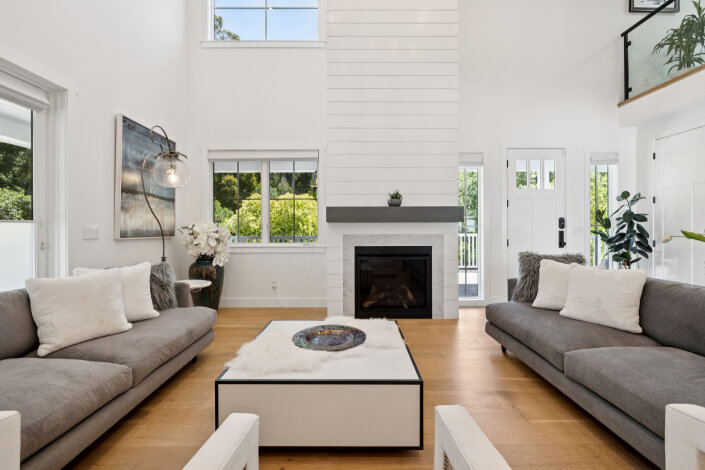Modern living room with high ceilings, two gray sofas with white pillows, a white coffee table, wooden floor, a black fireplace with a white shiplap wall, large windows, and green plants by the entrance.