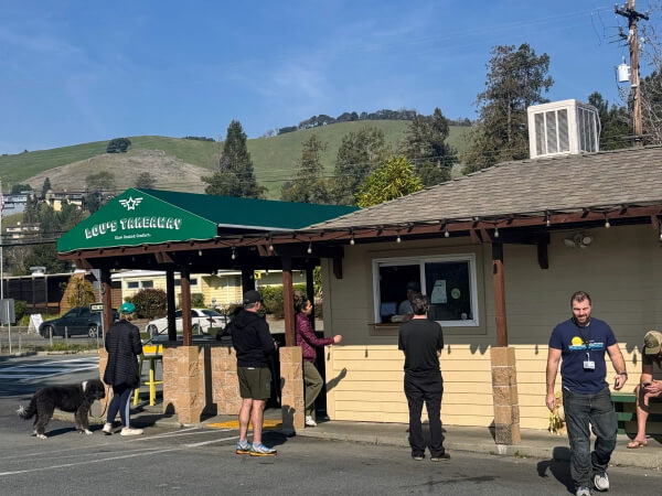 In Terra Linda San Rafael People stand and walk near a small takeout restaurant with a green awning reading “Lou’s Takeaway.” Hills and trees are visible in the background, while a large dog walks alongside one person in the parking lot.