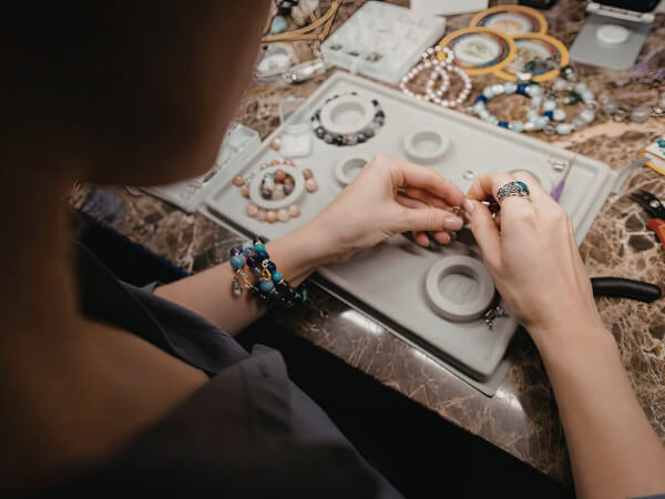 Marin estate sales separate valuables A person is making jewelry at a table, arranging beads and tools on a beige tray. Various finished jewelry pieces and supplies are scattered around on a marble surface.