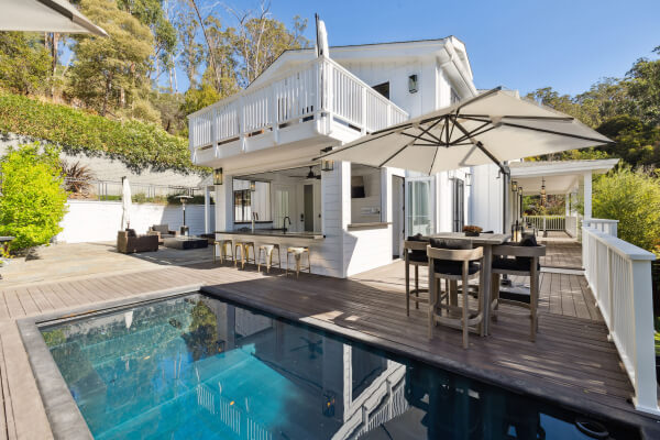Modern two-story white house with large windows, balcony, and outdoor living area featuring a pool, sun loungers, dining table with umbrella, and surrounded by greenery and trees.