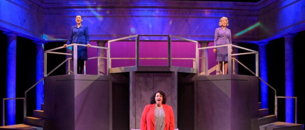 Marin Theater company in Mill Valley CA: Three women perform on stage; two stand on platforms in purple and blue outfits while one stands below in a red blazer, all under dramatic lighting with a minimal, modern set.