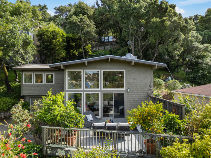 A modern gray house at 159 Terrace Avenue, San Rafael features large windows and a sloped roof among trees. The wooden deck offers outdoor seating and potted plants, while lush greenery and a garden create a tranquil setting.