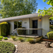 Charming single-story house at 1882 Mulberry Street in Yountville features light gray siding, large front windows, a black door with lit porch, and a small railed porch. Landscaped yard includes gravel, shrubs, and trees.
