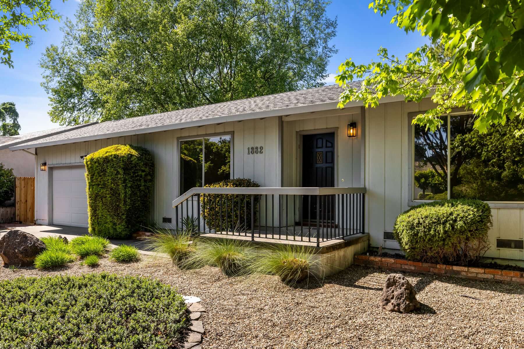 Charming single-story house at 1882 Mulberry Street in Yountville features light gray siding, large front windows, a black door with lit porch, and a small railed porch. Landscaped yard includes gravel, shrubs, and trees.