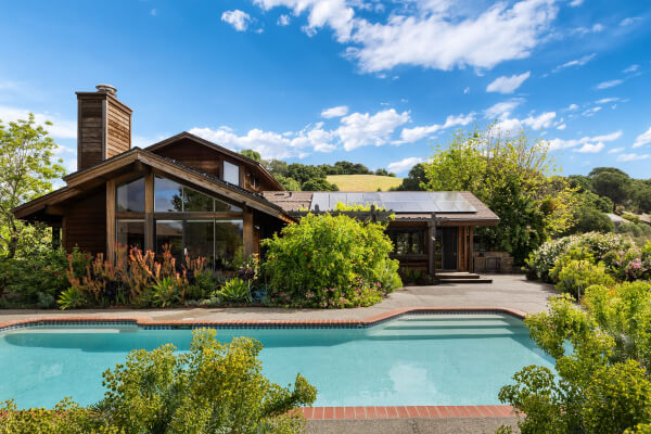 95 Irving Drive in San Anselmo is a wooden house with large windows and solar panels, set behind a swimming pool and surrounded by lush greenery under a bright blue sky with scattered clouds.