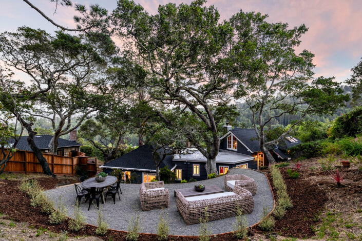 Outdoor patio with wicker furniture and dining table among trees, facing a modern house with large windows on a hillside at sunset.