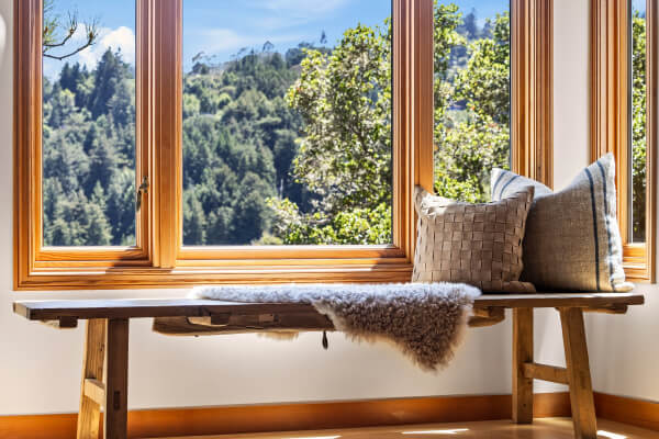 A wooden bench with a sheepskin throw and two pillows by broad windows overlooks green trees and mountains beneath a blue sky.