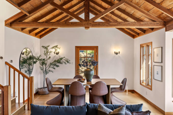 Dining room featuring exposed wooden beams, a wood table, brown chairs, large vase centerpiece, round mirror, plant and big windows.