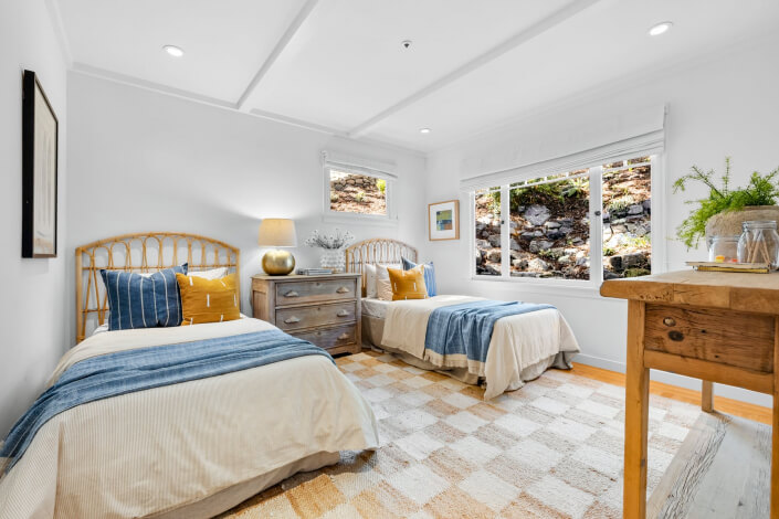 A sunlit bedroom with two single beds featuring blue and yellow pillows, a wooden nightstand, large windows, and a rustic console table with plants.