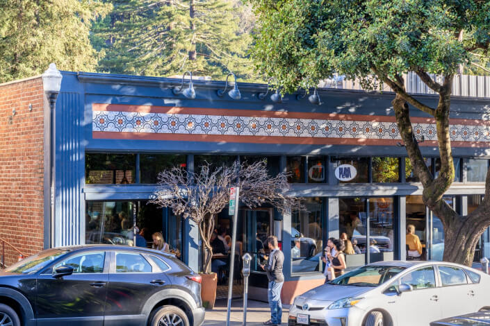 People gather outside Playa Mill Valley with large windows and geometric tile decor, parked cars and trees nearby on a sunny day.