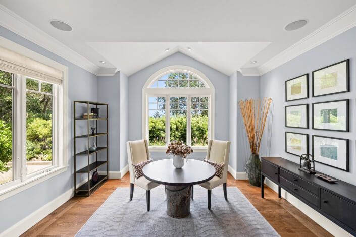 Sunlit room featuring a large arched window, two white chairs around a round table, black shelving, a sideboard adorned with decorative items, framed artwork on the wall, and wooden floors partially covered by a light gray rug.