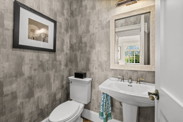 A compact bathroom featuring gray painted walls, a toilet, a white pedestal sink, and a mirror above the sink. A towel with a blue pattern hangs to one side, accompanied by a wall-mounted light and a framed photograph on the wall. The reflection of a window is visible in the mirror.