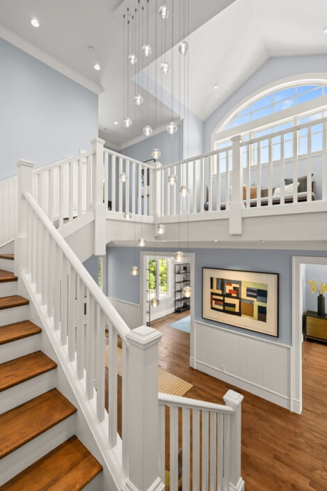 Modern home interior featuring a white staircase with wooden steps, high ceilings, expansive windows, glass globe pendant lights, and abstract wall art. Bright natural light streams into the open and airy space.