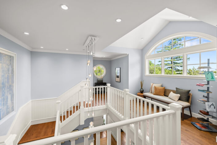 Bright upstairs landing with white railings, wood floors, a modern chandelier, large arched window, built-in bench with pillows, and shelves filled with books. Blue walls and views of nature contribute to the airy and inviting atmosphere.