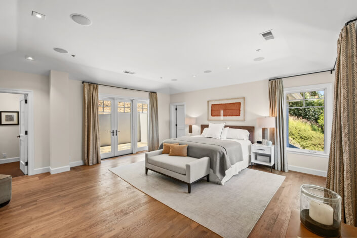 Bright, spacious bedroom with a large bed, bench, and nightstands. Hardwood floors, windows with curtains, glass doors to a patio, and neutral decor create a modern atmosphere.