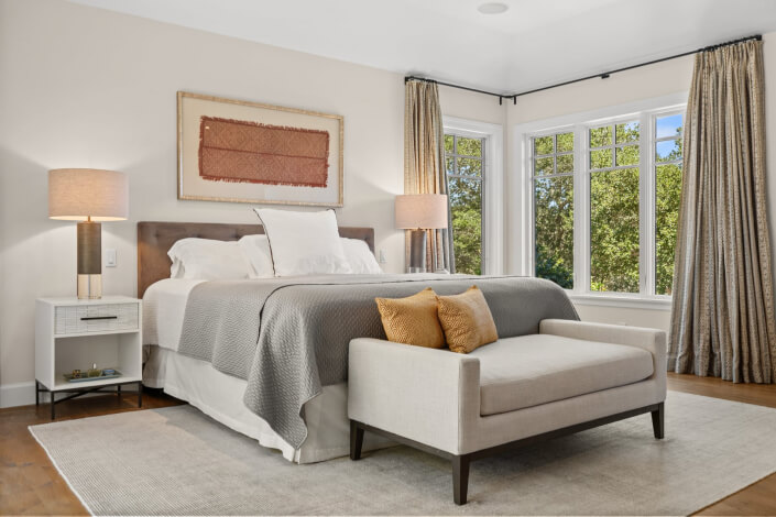 Modern bedroom featuring a neatly made bed with gray bedding, two bedside tables each topped with a lamp, a framed artwork displayed above the bed, a cream-colored bench at the foot of the bed, large windows draped with curtains, and a beige rug laid over wooden flooring.