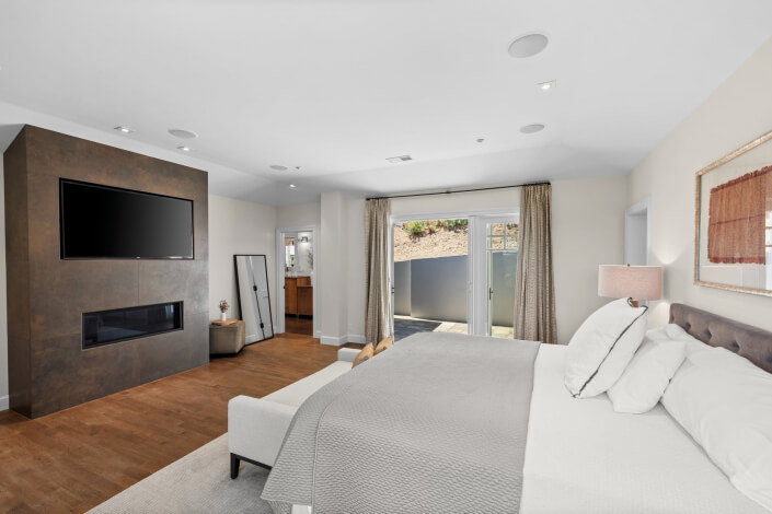 Modern bedroom featuring a large bed with neutral bedding, a wall-mounted TV above the fireplace, wooden floors, a floor mirror, and glass doors opening to the outdoors. Natural light fills the room.